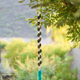 Large gray-green oval stone beads strung with clear glass lamp worked beads, hanging in two strands vertically; anchored by two pieces of teal and white fused glass, against blurred landscape background.