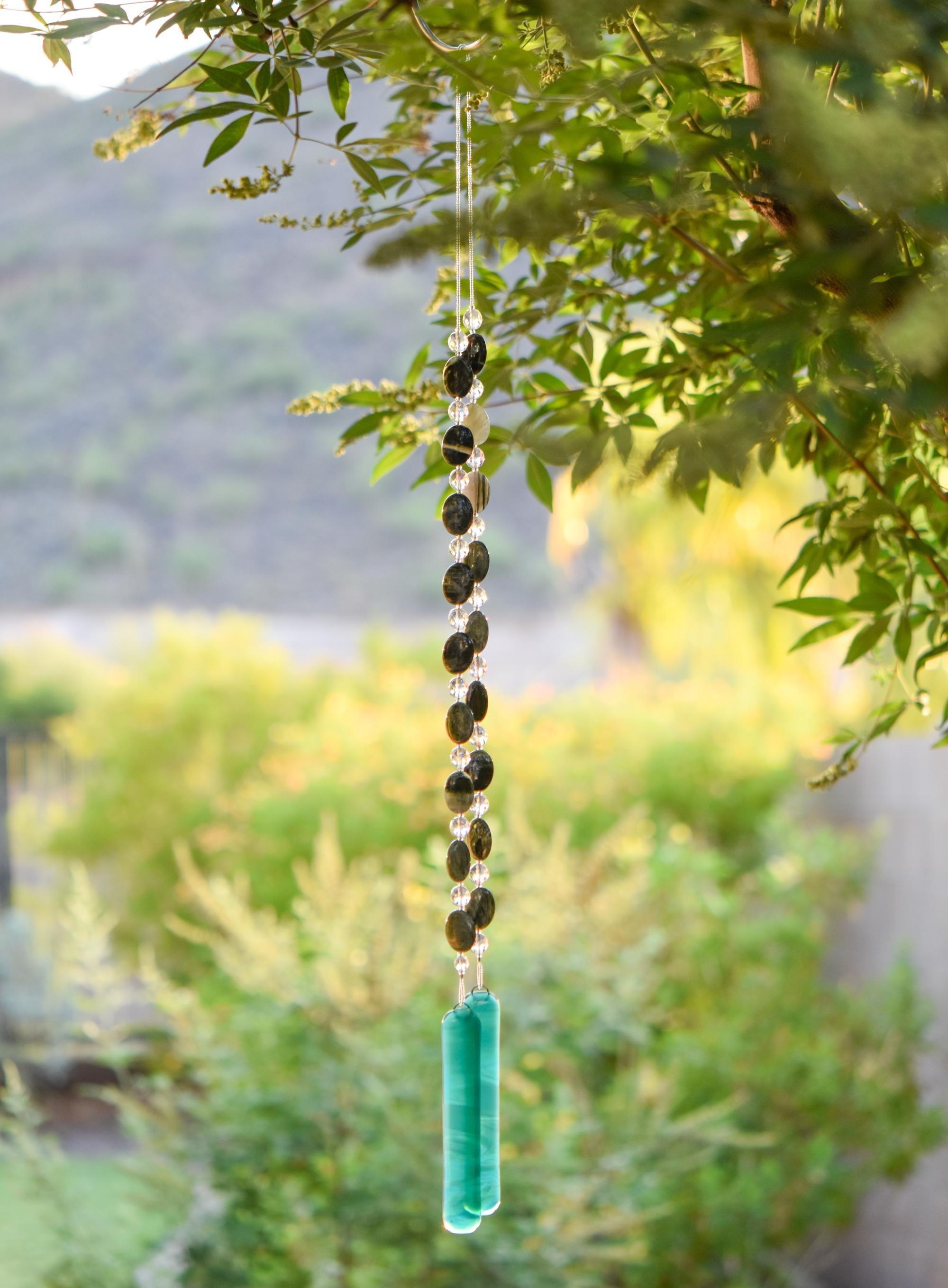 Large gray-green oval stone beads strung with clear glass lamp worked beads, hanging in two strands vertically; anchored by two pieces of teal and white fused glass, against blurred landscape background.