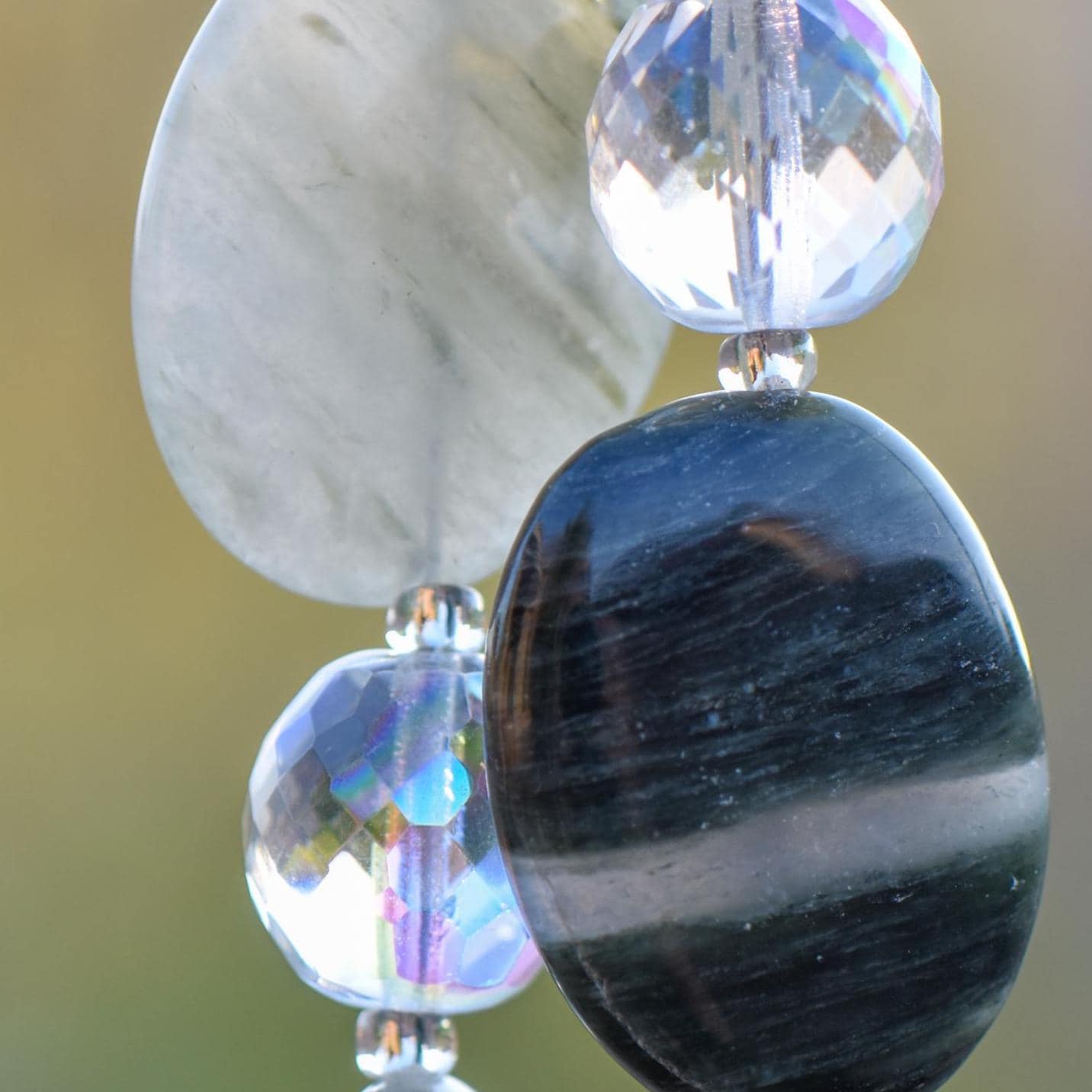 Close-up of two beaded decorative strands featuring various polished seaweed quartz stones and faceted crystal beads. The stones are oval and teardrop-shaped, in shades of gray, black, and green. The crystal beads are round, clear and faceted, reflecting light. The strands are suspended against a blurred, neutral background.