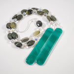 Large gray-green oval stone beads strung with clear glass lamp worked beads, hanging in two strands vertically; anchored by two pieces of teal and white fused glass, coiled on a white surface.