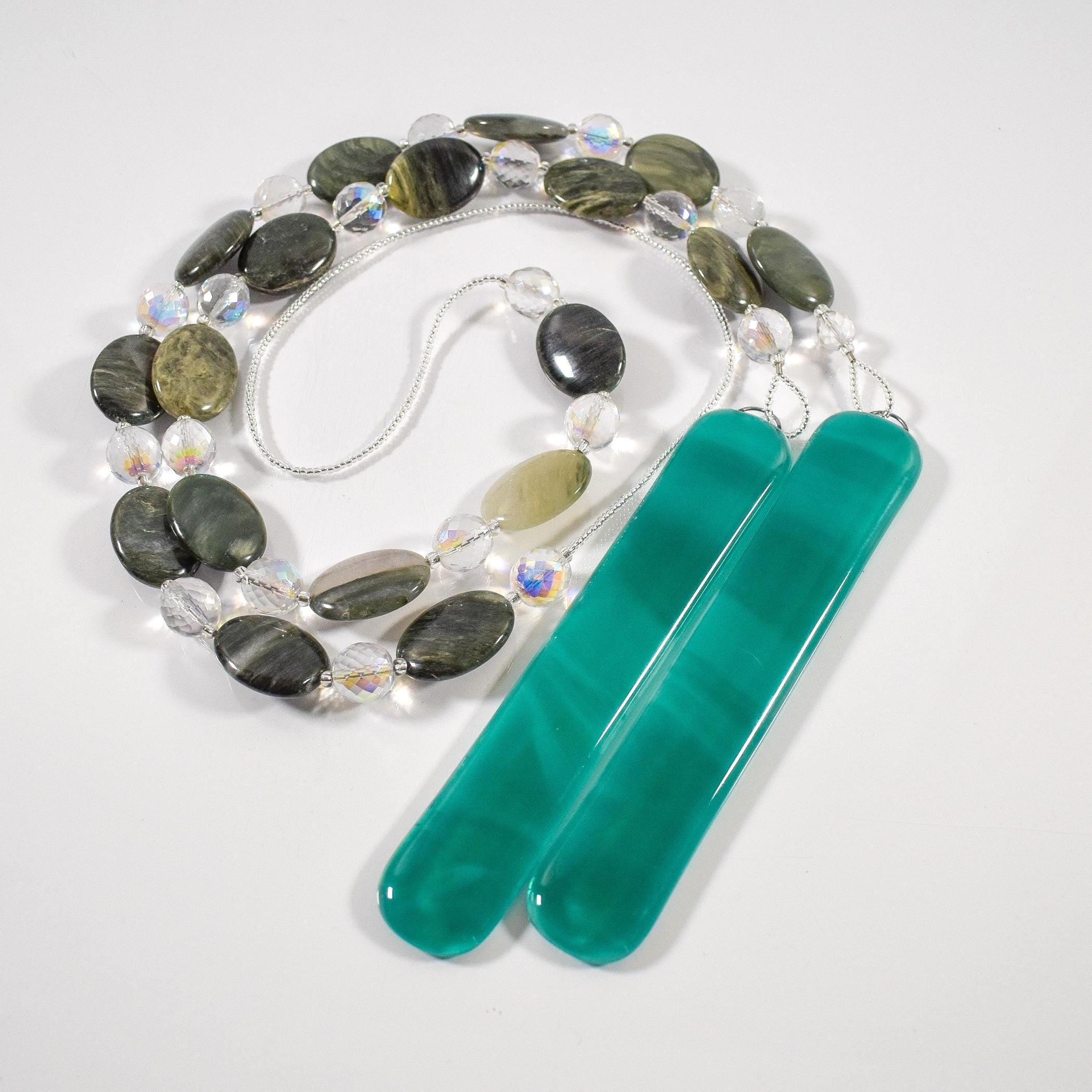 Large gray-green oval stone beads strung with clear glass lamp worked beads, hanging in two strands vertically; anchored by two pieces of teal and white fused glass, coiled on a white surface.