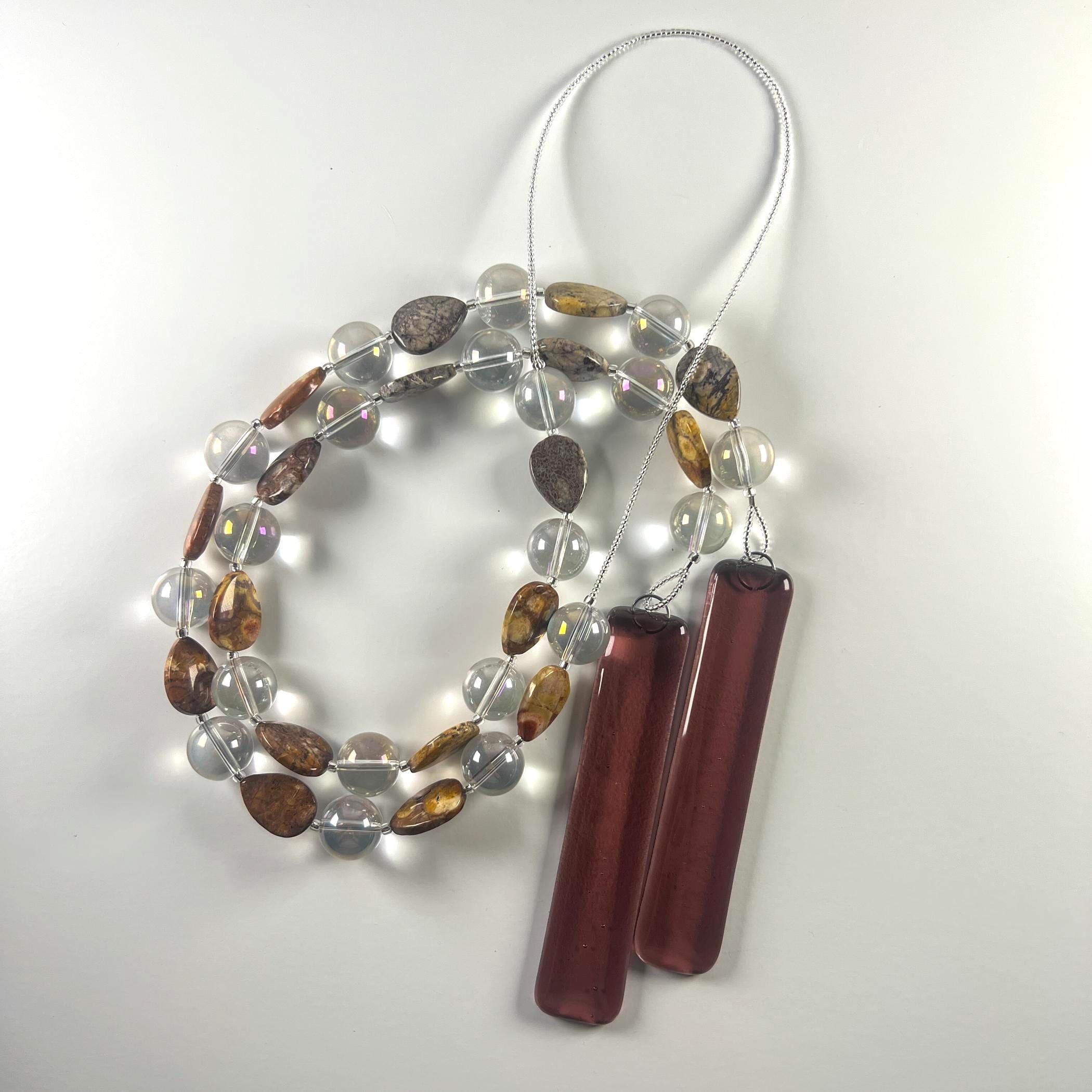 Cinjo Handmade wind chime coiled on a white surface, clearly showing the beaded wire and the two piece of fused glass in pale purple. The Rhyolite stone beads are tear drop shaped and there is a larger, clear, reflective glass bead in between each stone bead. The stone beads are marked and in shades of brown, tan and gray. Any wire not covered by larger beads are covered with silver-lined seed beads.