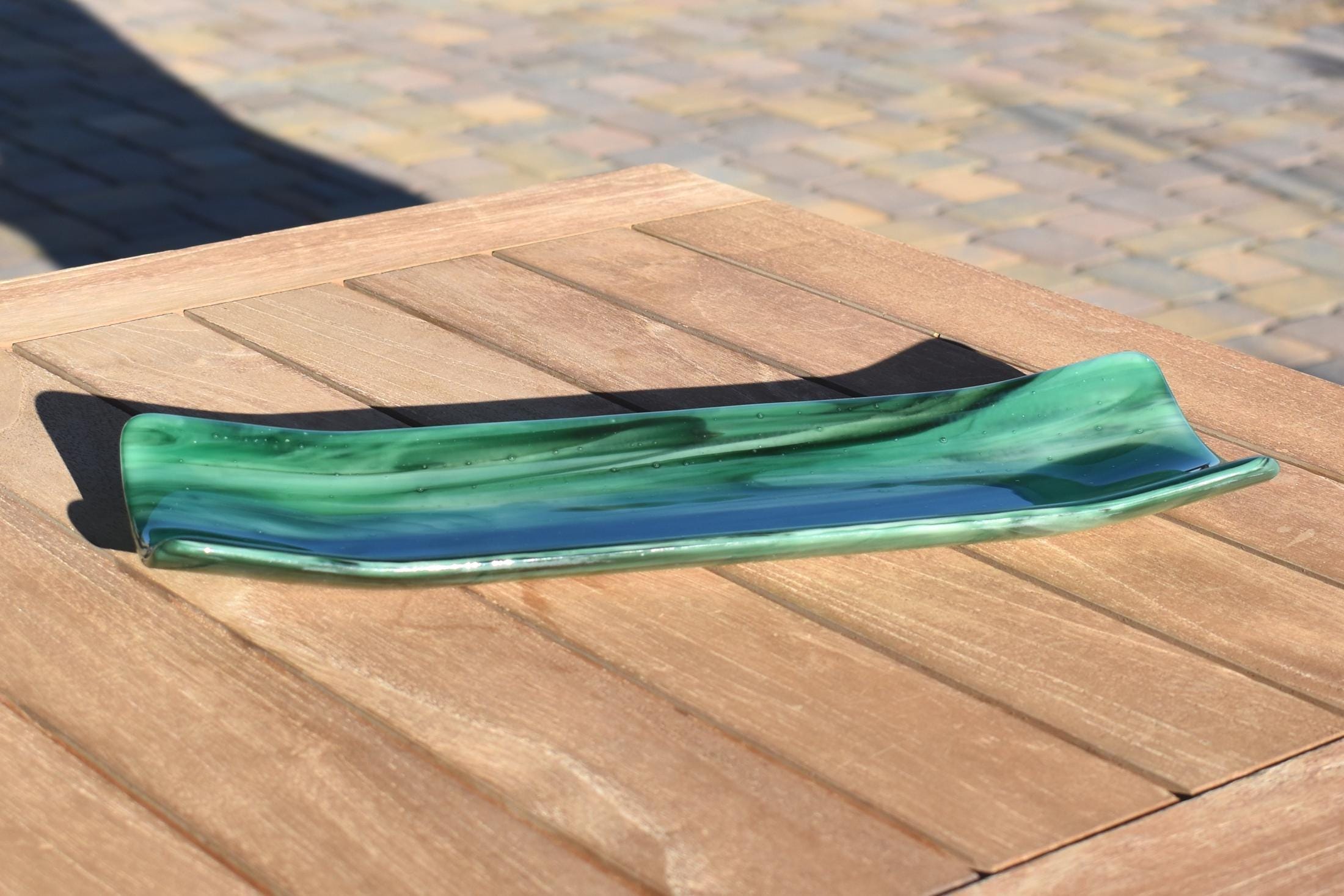 Green fused glass channel tray on an outdoor slatted wood table in sunlight, highlighting the glossy surface and color shifts.