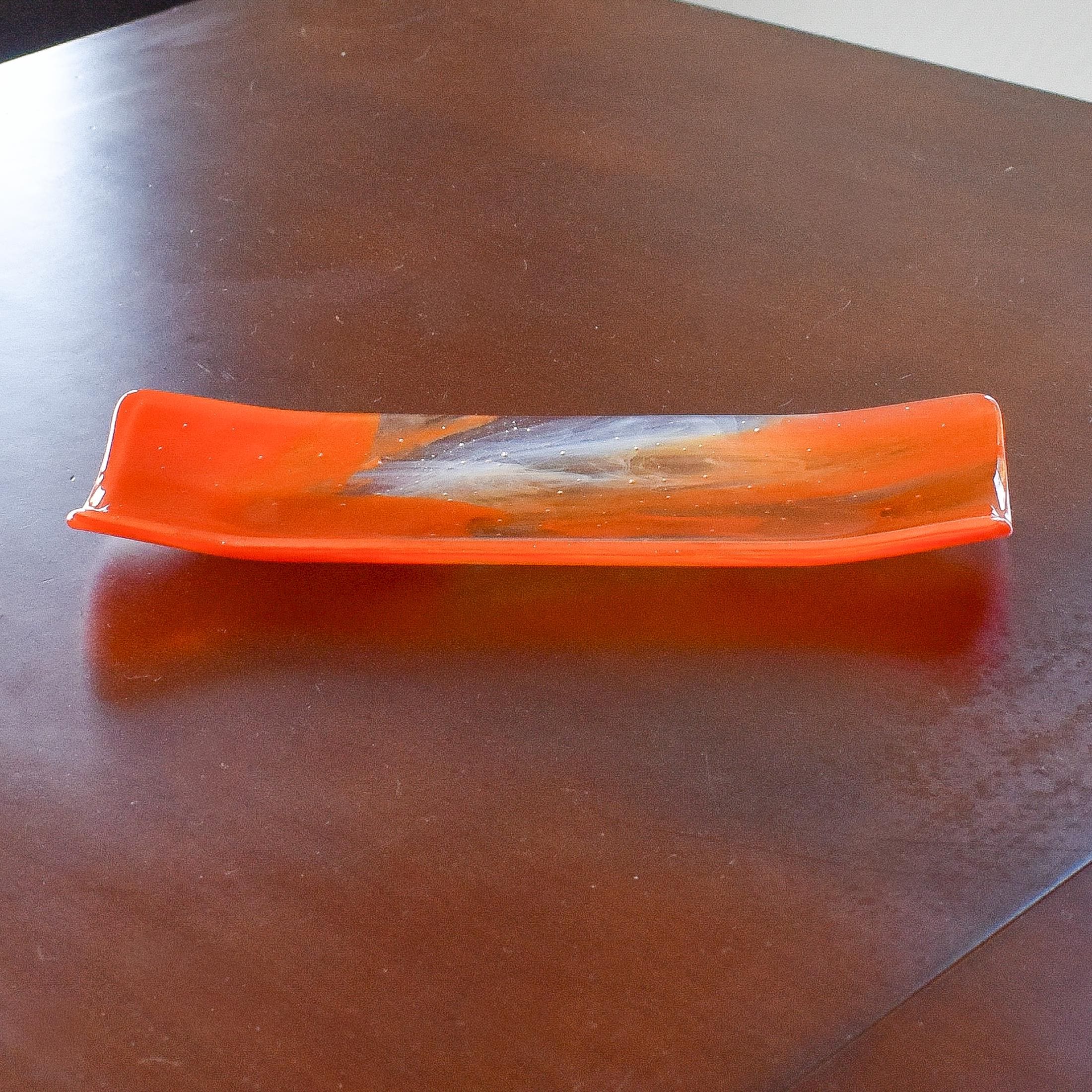 Orange and clear fused glass channel tray photographed from the side on a brown wood table.