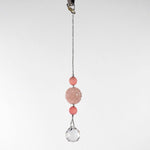 Large round carved rose quartz bead flanked by two smaller pink stone beads and anchored by round crystal prism, hanging form a hook in front of white background.