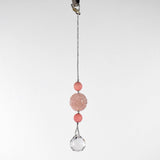 Large round carved rose quartz bead flanked by two smaller pink stone beads and anchored by round crystal prism, hanging form a hook in front of white background.