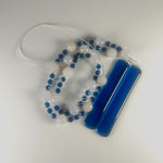 A coiled wind chime with two strings of white and blue beads, and two pieces of dark blue glass, coiled on a gray surface. The beads are interspersed with small blue beads.