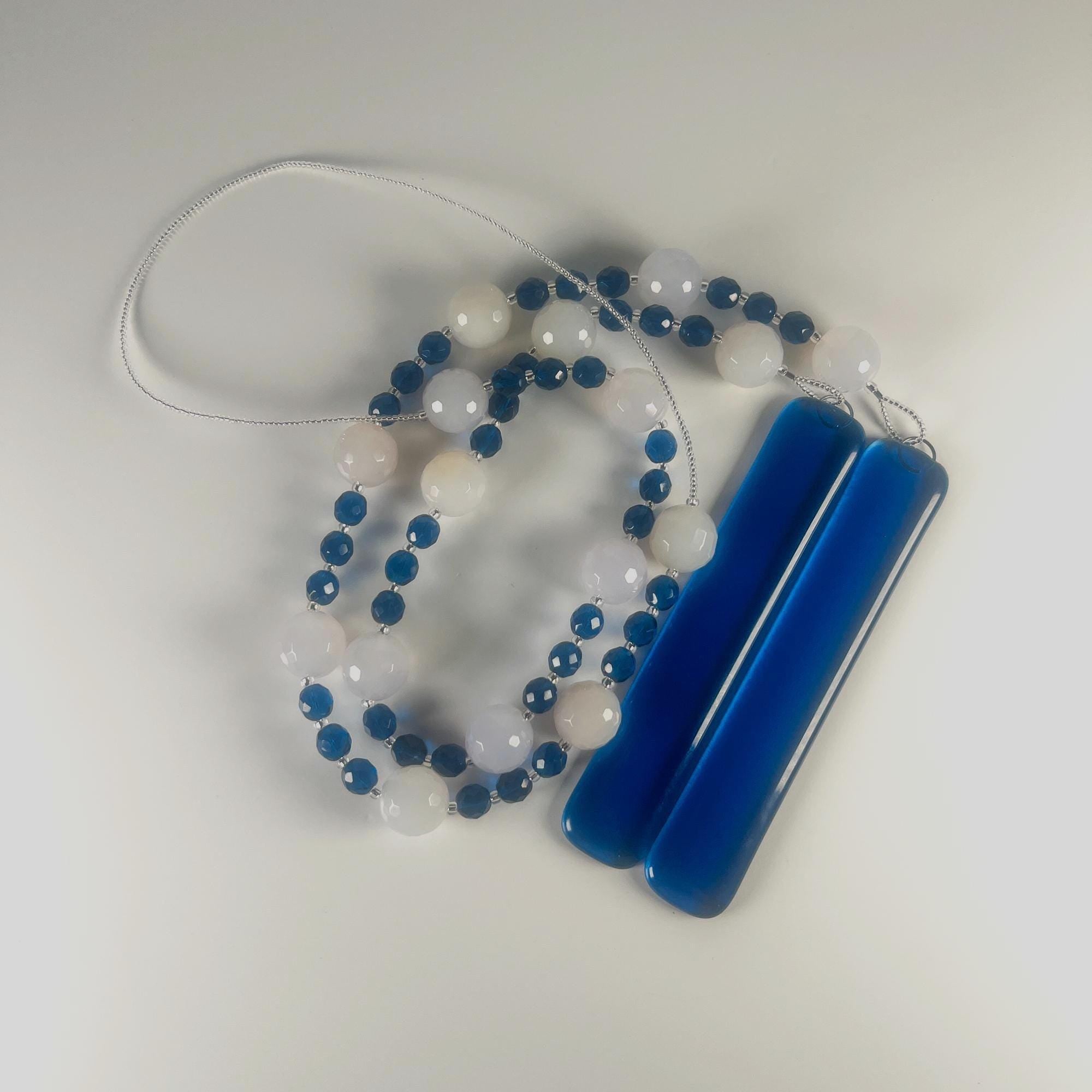 A coiled wind chime with two strings of white and blue beads, and two pieces of dark blue glass, coiled on a gray surface. The beads are interspersed with small blue beads.