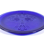A cobalt blue glass plate with a raised cross design in the center. The plate is round with a slightly raised edge. The cross is detailed with floral-like embellishments.