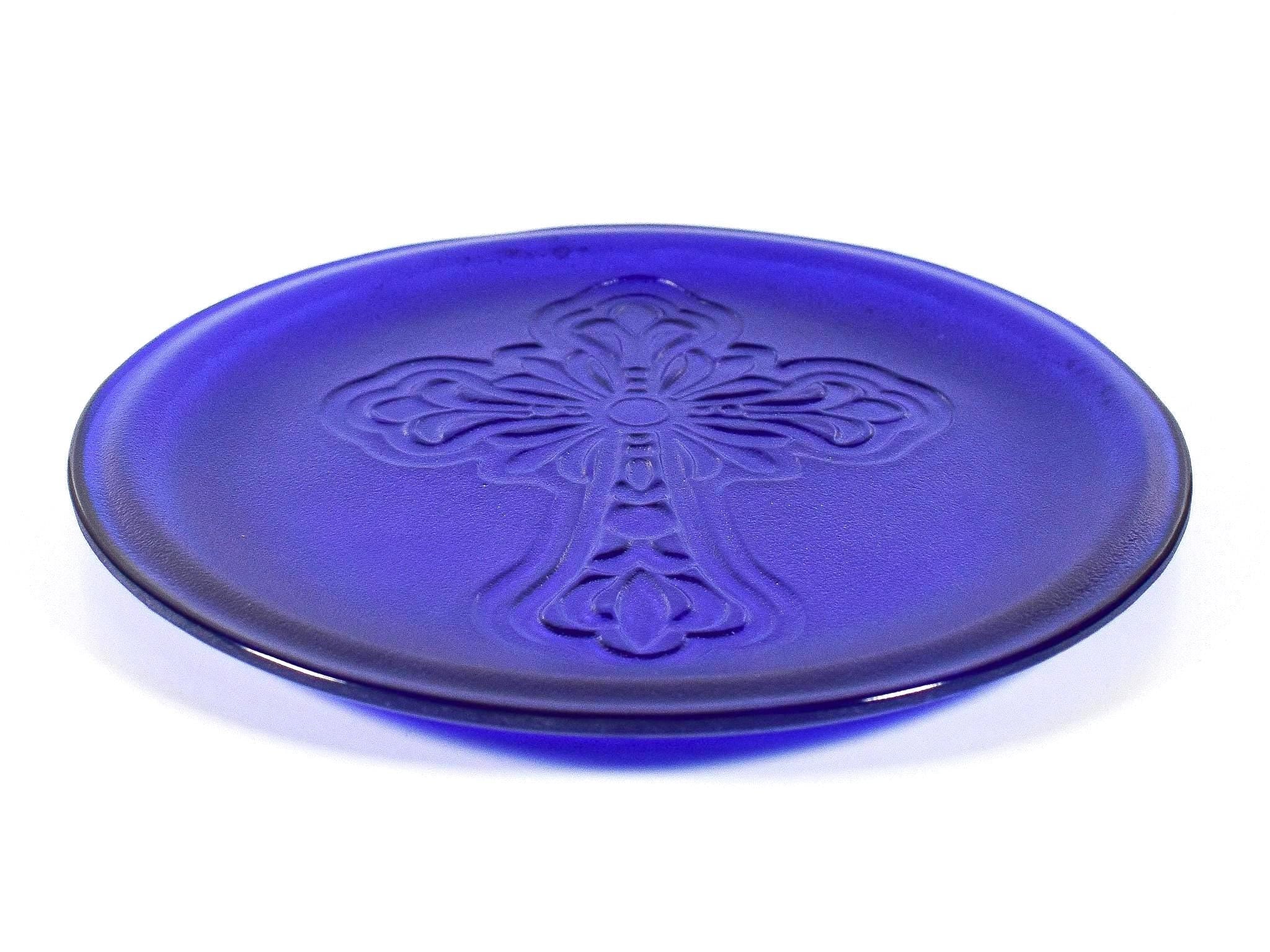 A cobalt blue glass plate with a raised cross design in the center. The plate is round with a slightly raised edge. The cross is detailed with floral-like embellishments.