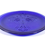 A cobalt blue glass plate with a raised cross design in the center. The plate is round with a slightly raised edge. The cross is detailed with floral-like embellishments.