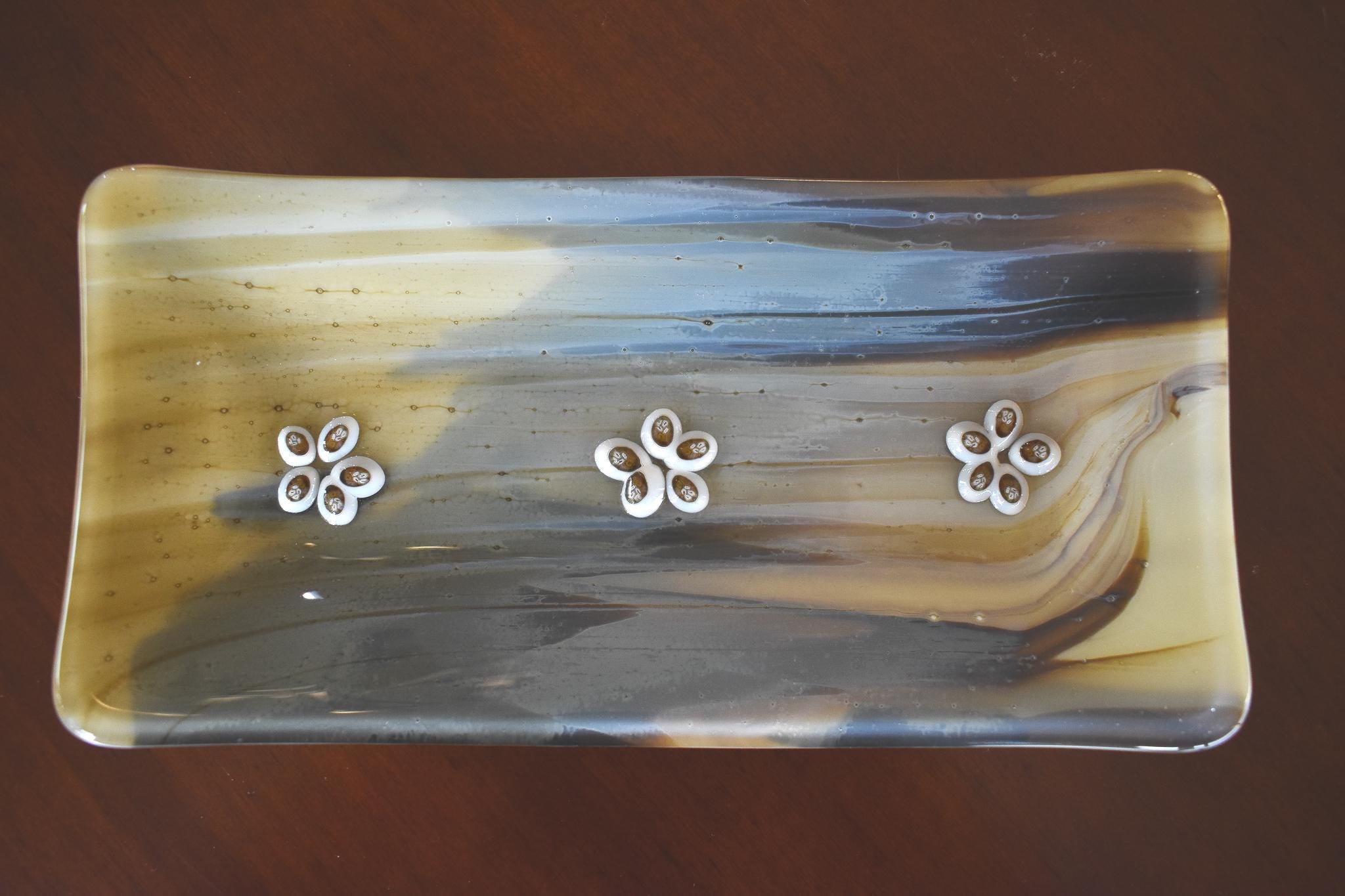 A rectangular glass dish with a marbled pattern in shades of brown, tan, and gray and gold. Three white flower-shaped embellishments are evenly spaced across the top. The dish is likely used for decorative purposes or as a small serving tray.