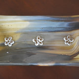 A rectangular glass dish with a marbled pattern in shades of brown, tan, and gray and gold. Three white flower-shaped embellishments are evenly spaced across the top. The dish is likely used for decorative purposes or as a small serving tray.