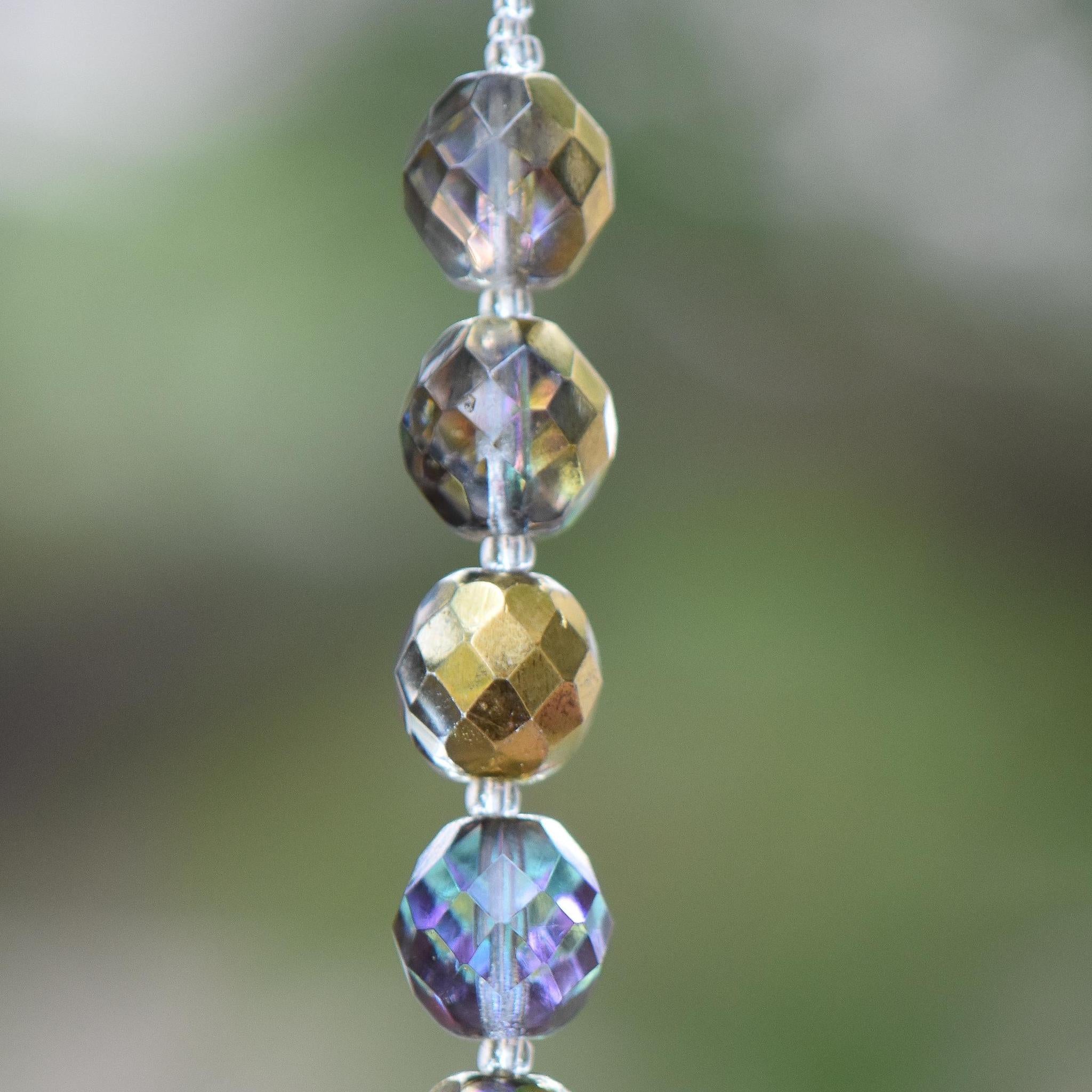 Close-up of a strand of faceted beads in shades of gold, brown, and iridescent purple with iridescence. The beads are strung together with a thin, clear string, creating a shimmering effect. The background is a soft, blurred green.