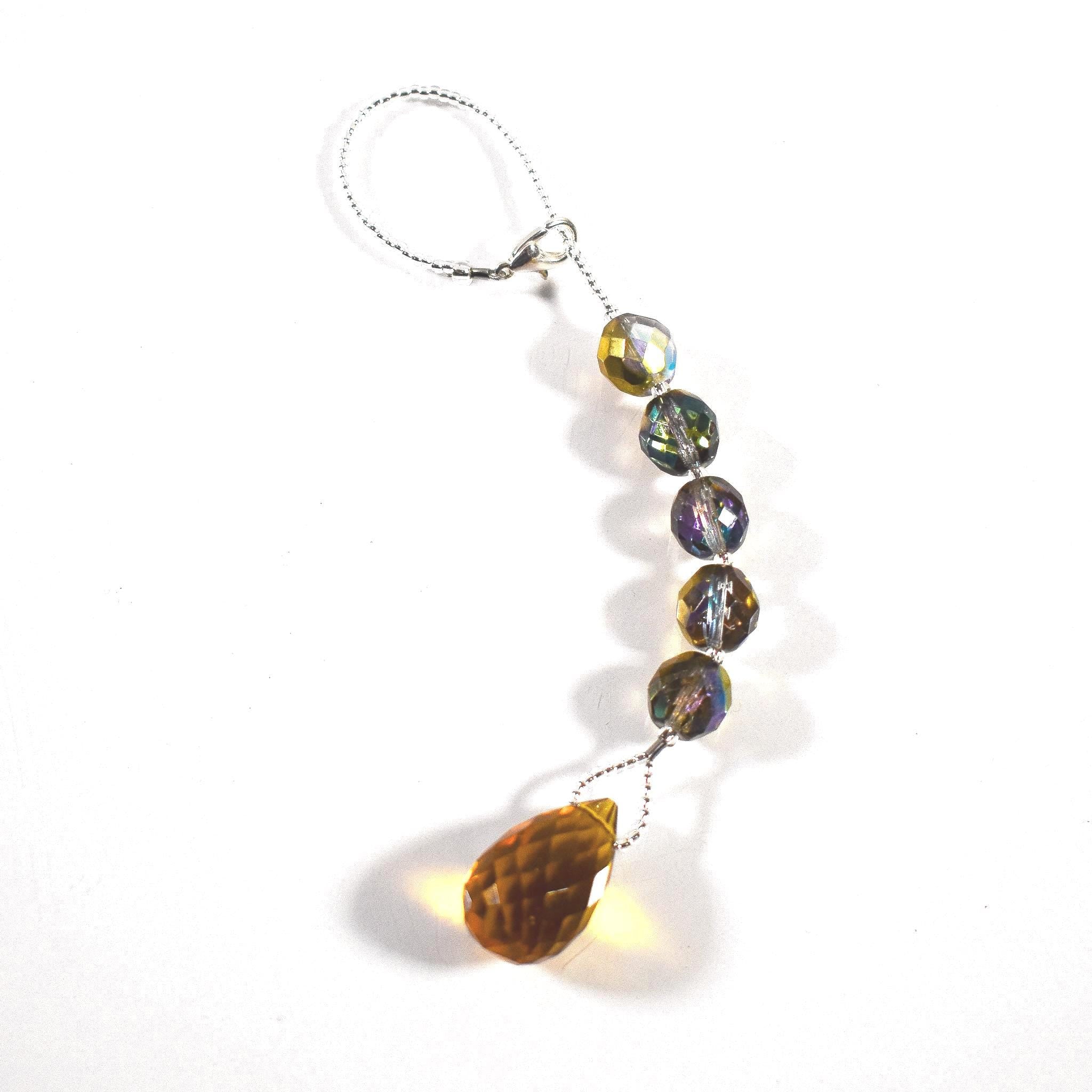 A decorative beaded ornament with a silver loop and clasp. The ornament features a string of iridescent, faceted beads in shades of purple, green, and gold, leading to a large, amber-colored teardrop-shaped crystal.