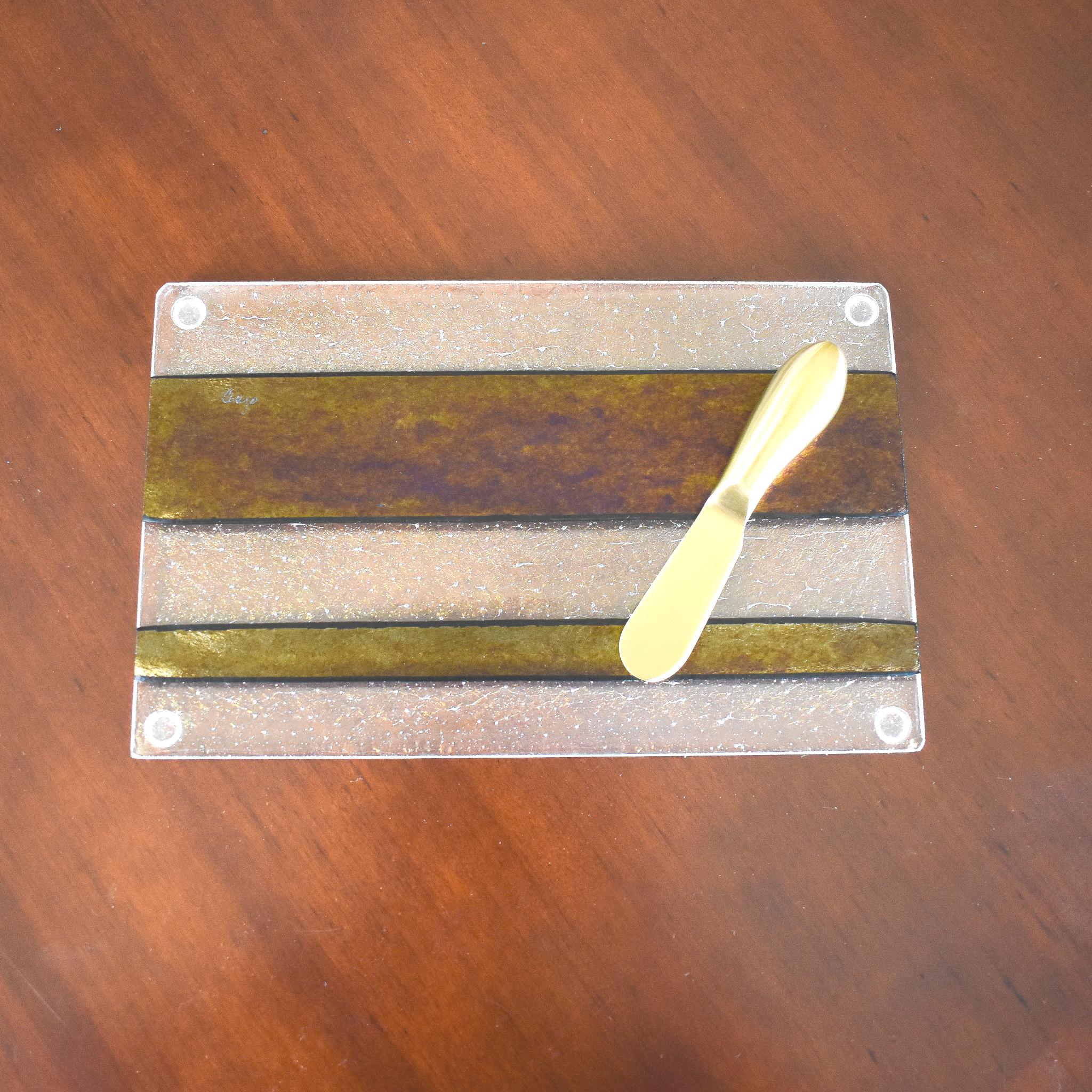 A clear glass serving board with a textured surface and gold and black stripes. A gold butter knife is on the board.