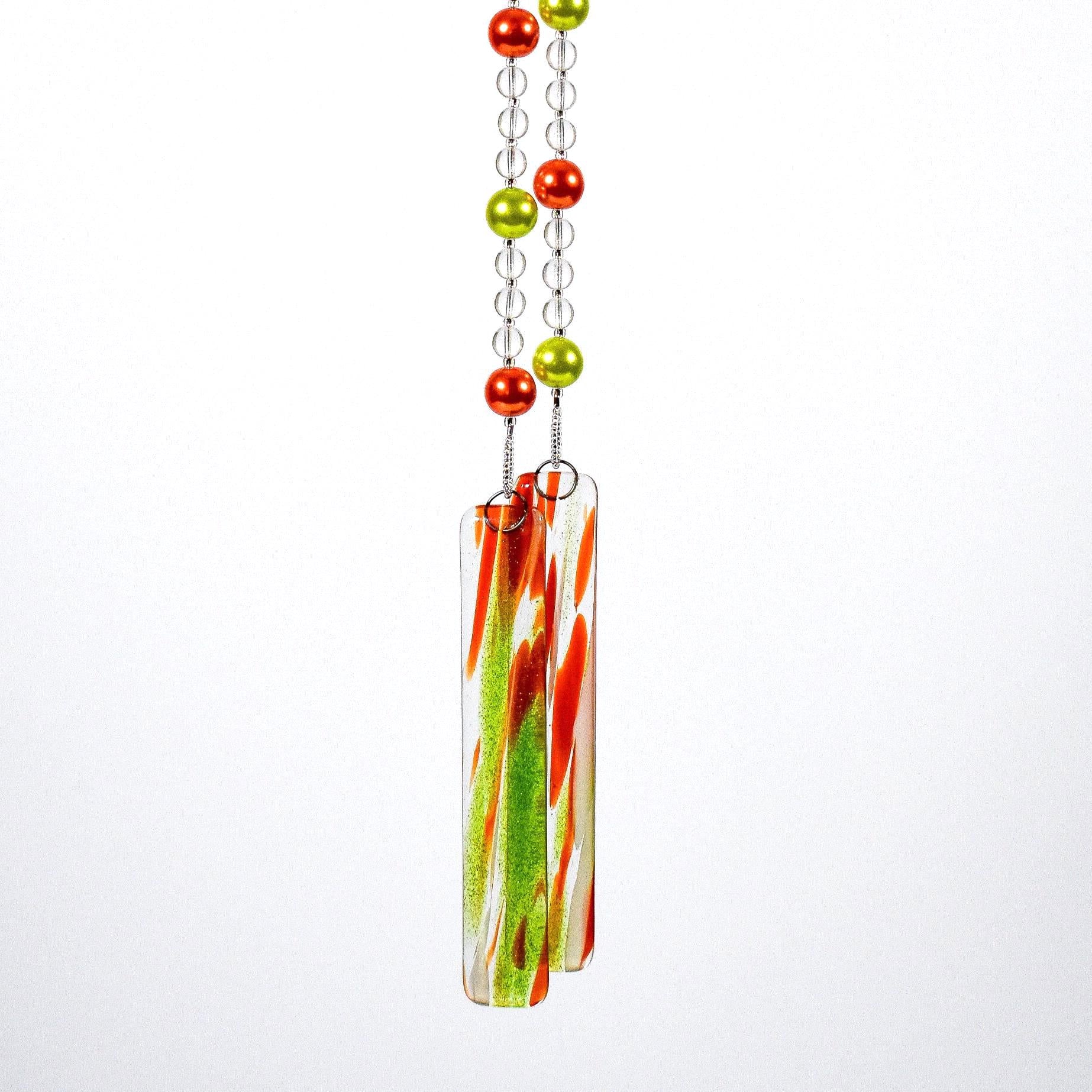Two strings of glass beads hanging against white background, anchored by two pieces of green and orange fused glass.
