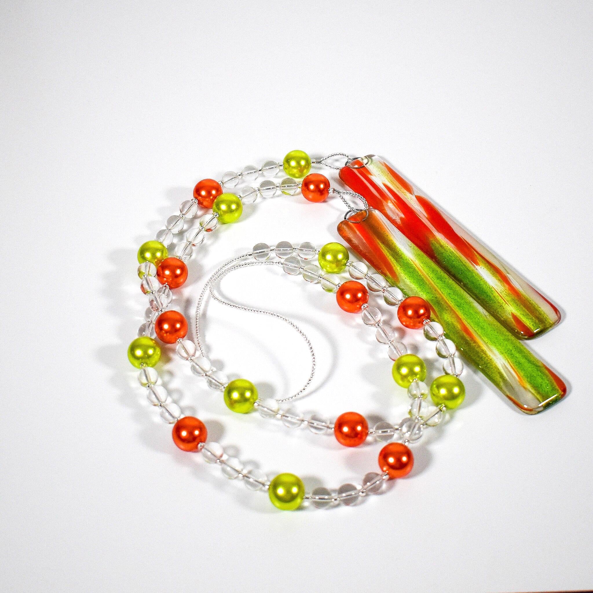 LIme green and bright orange pearl beads strung with smaller clear beads coiled on a white surface. Both ends of the wire are connected to rectangular pieces of fused glass with sparkling green and orange streaks.