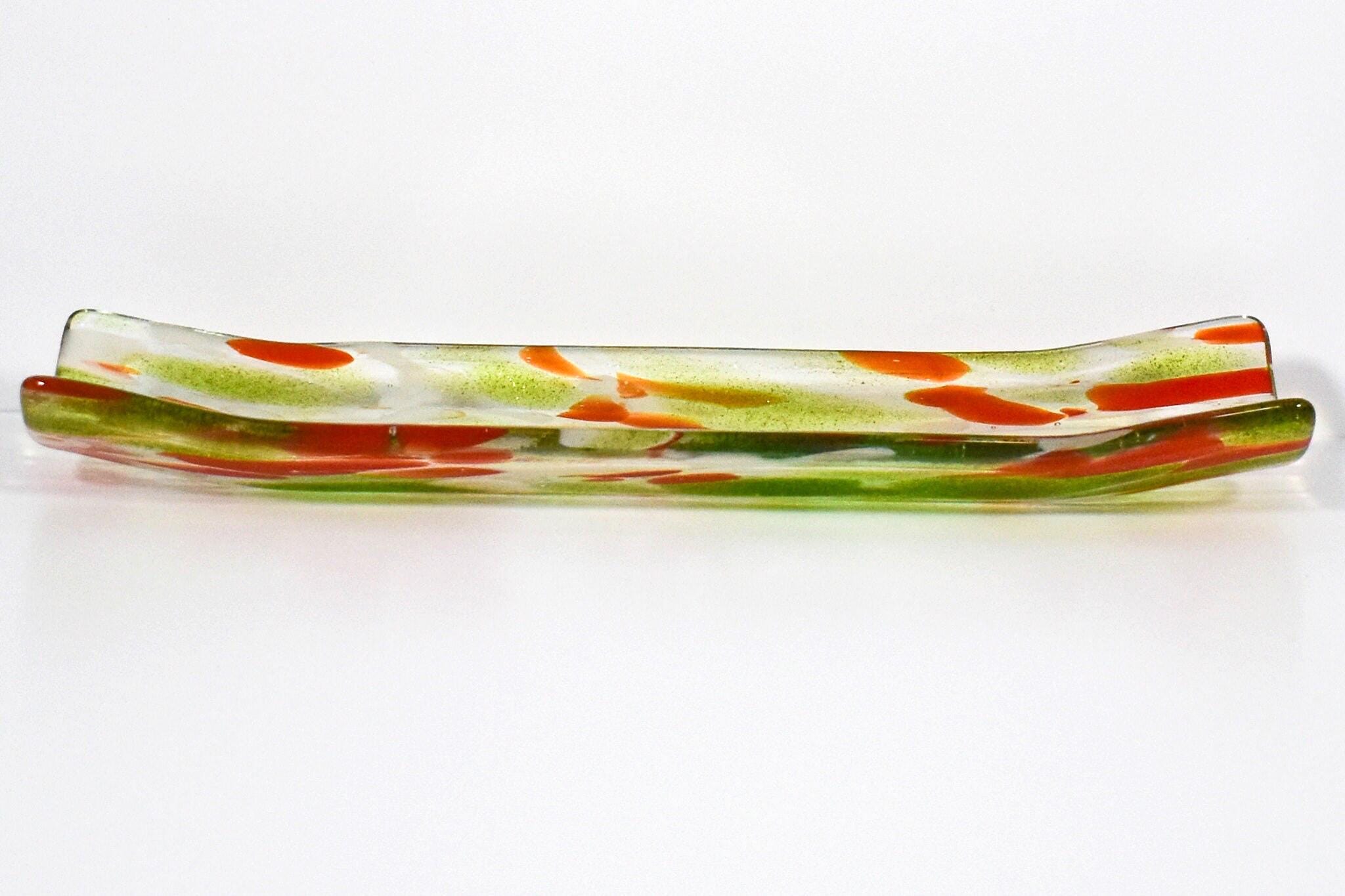 A long, rectangular glass dish with a curved shape. The dish is clear with green and orange color accents. The green is in the form of streaks and the orange is in the form of small, irregular shapes. The dish is likely used for serving or display purposes.