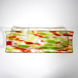 A rectangular glass dish with a decorative pattern. The dish is clear with a speckled design of green and orange shapes. The colors are distributed across the surface, creating a textured appearance. The dish has slightly curved edges and sits on a white surface.
