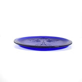 A round, cobalt blue plate with a raised cross design in the center. The plate has a smooth, glossy finish and a slightly raised edge. The plate is made of glass.