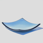 Denim blue square glass dish with black trim and slightly sloped corners, viewed from the side in front of a white background.