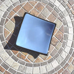 A square, light blue glass dish with a black rim. The dish is sitting on a circular stone patio.