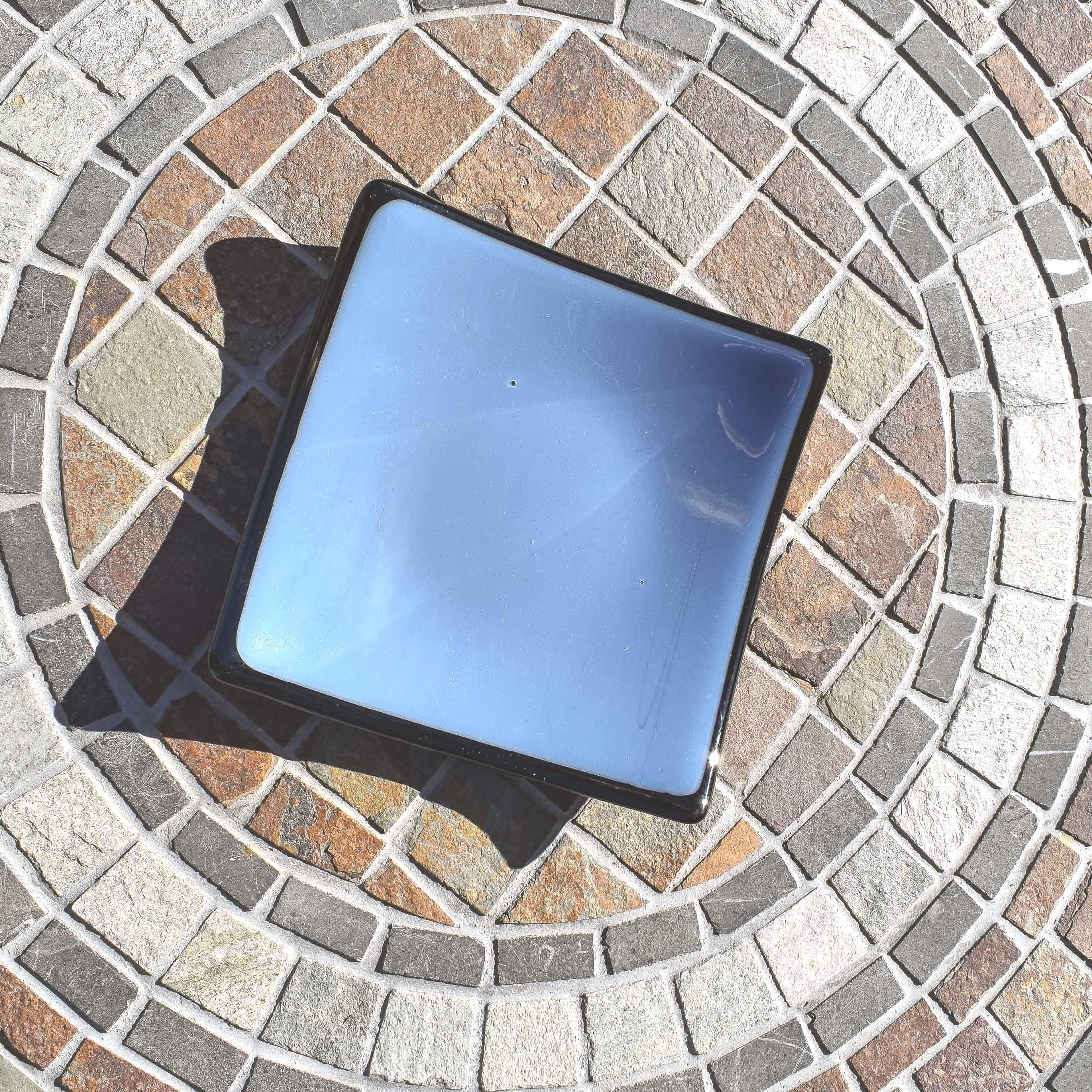 A square, light blue glass dish with a black rim. The dish is sitting on a circular stone patio.