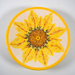 A white glass bowl with a yellow rim features a hand-painted sunflower design. The sunflower is made up of yellow, orange, brown, and green dots.