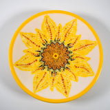 A white glass bowl with a yellow rim features a hand-painted sunflower design. The sunflower is made up of yellow, orange, brown, and green dots.