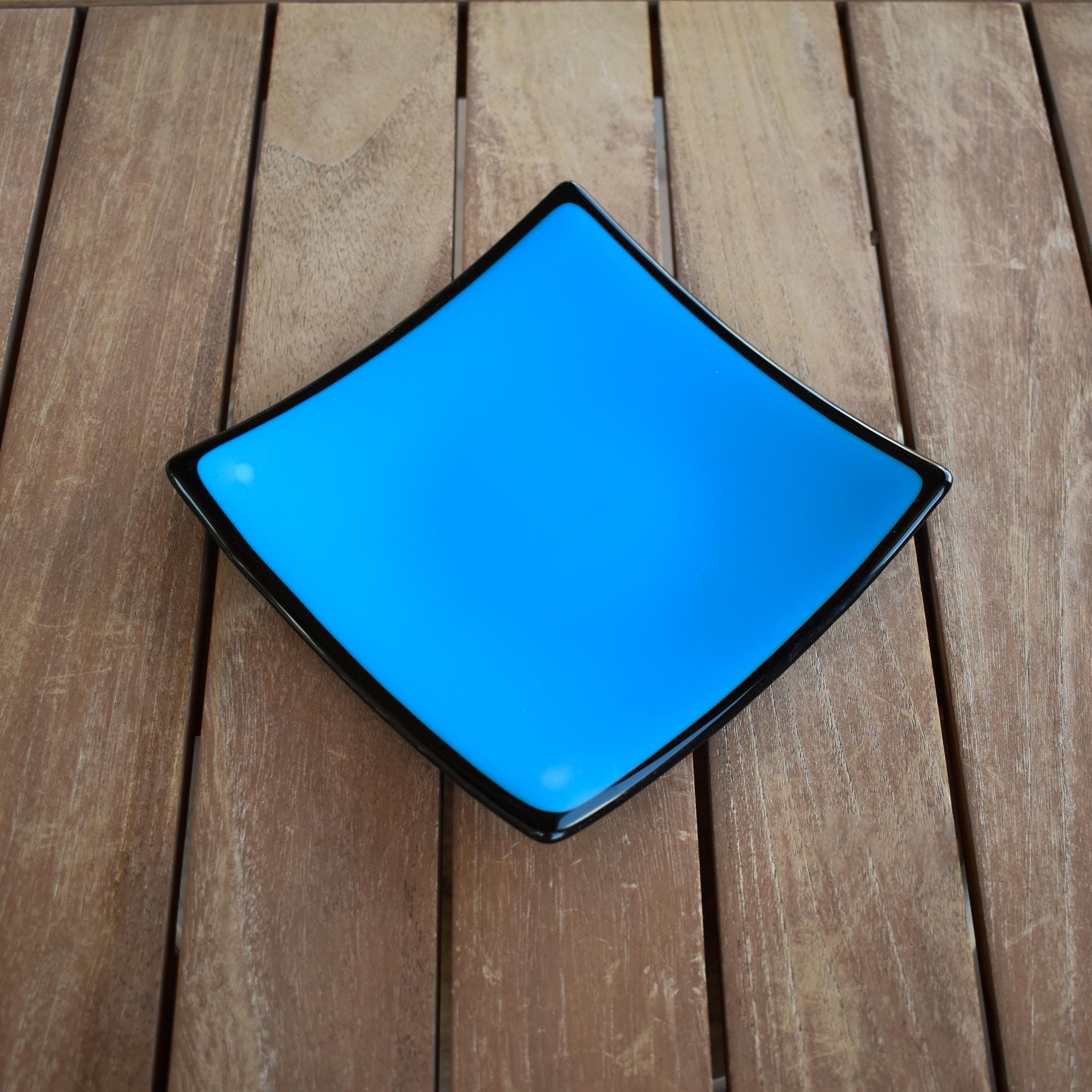 Blue, square fused glass tray with black back and trim, sitting on a wood table.