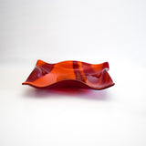 Large square glass bowl with wavy edges and a square center foot, viewed from the side, sitting on a white surface.