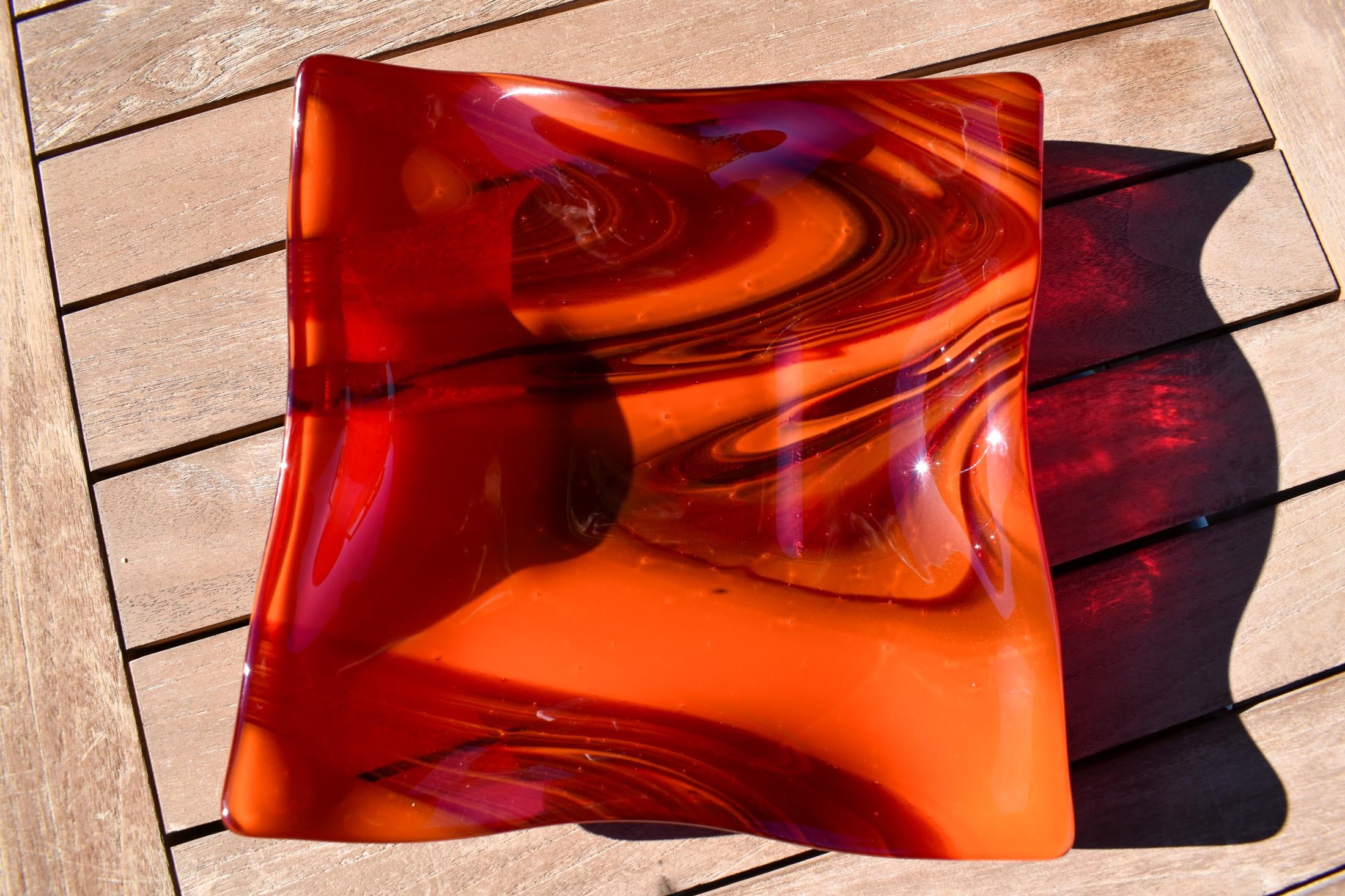 Large square glass bowl with wavy edges and a square center foot, viewed from above sitting on a slatted wood table, outside in sun light.