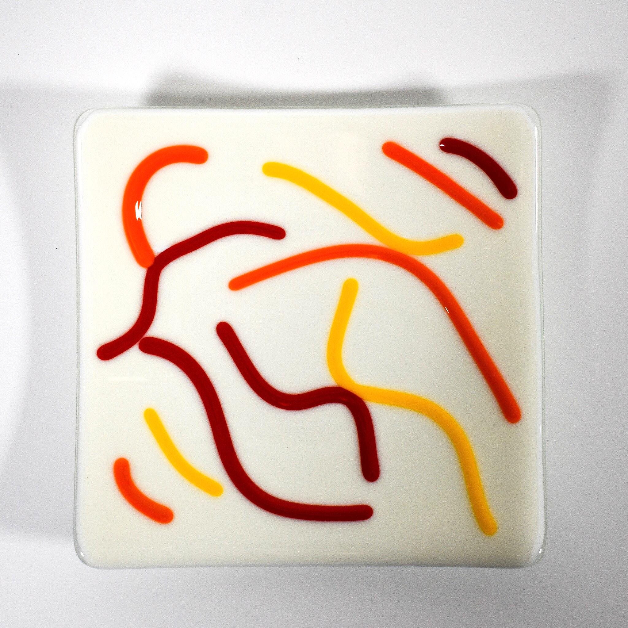 A creamy white square glass plate with a design of red, orange, and yellow lines, viewed from above, on white surface.