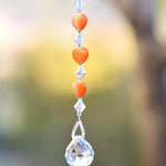 A crystal suncatcher with a clear crystal ball at the bottom and a string of orange heart-shaped beads above it.