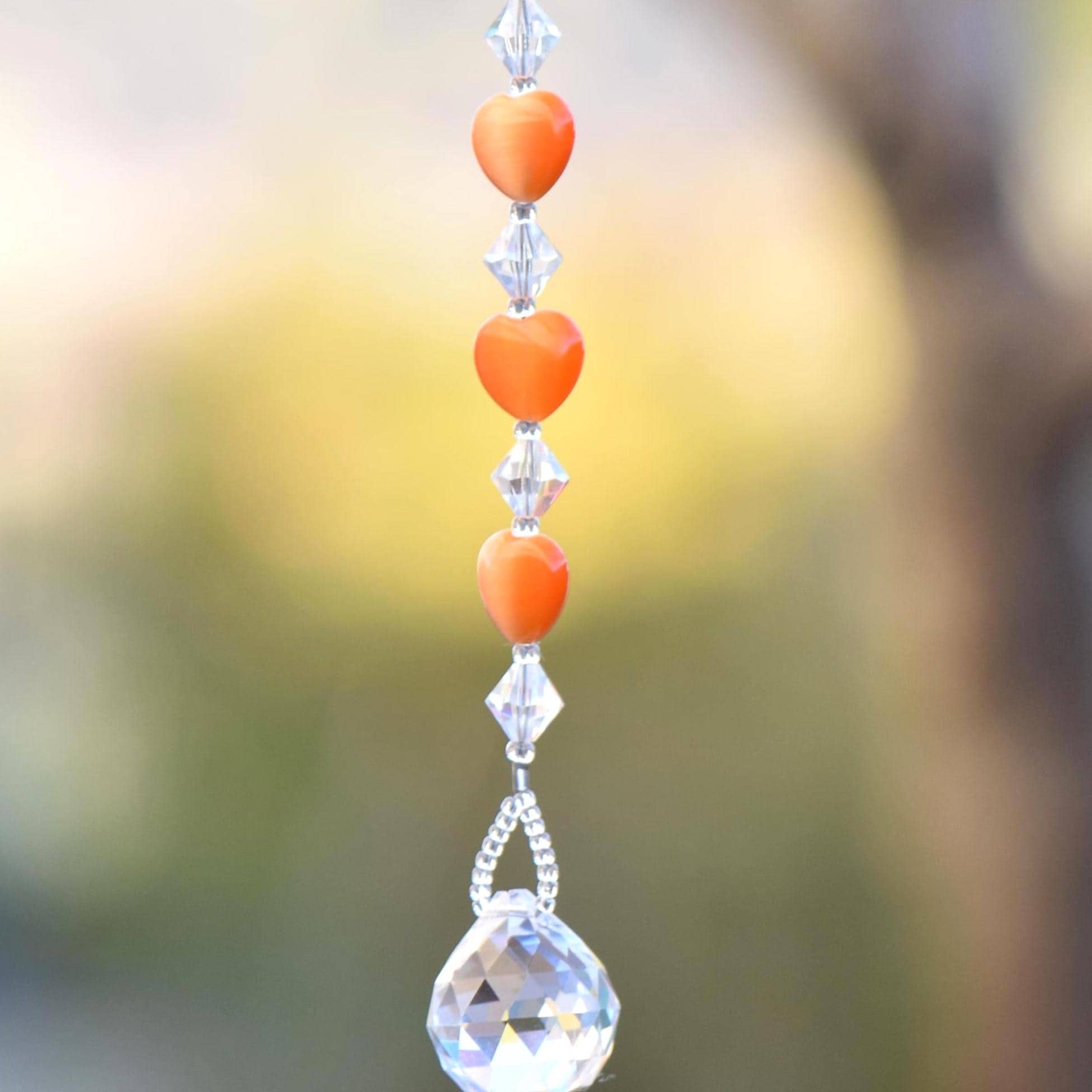 A crystal suncatcher with a clear crystal ball at the bottom and a string of orange heart-shaped beads above it.