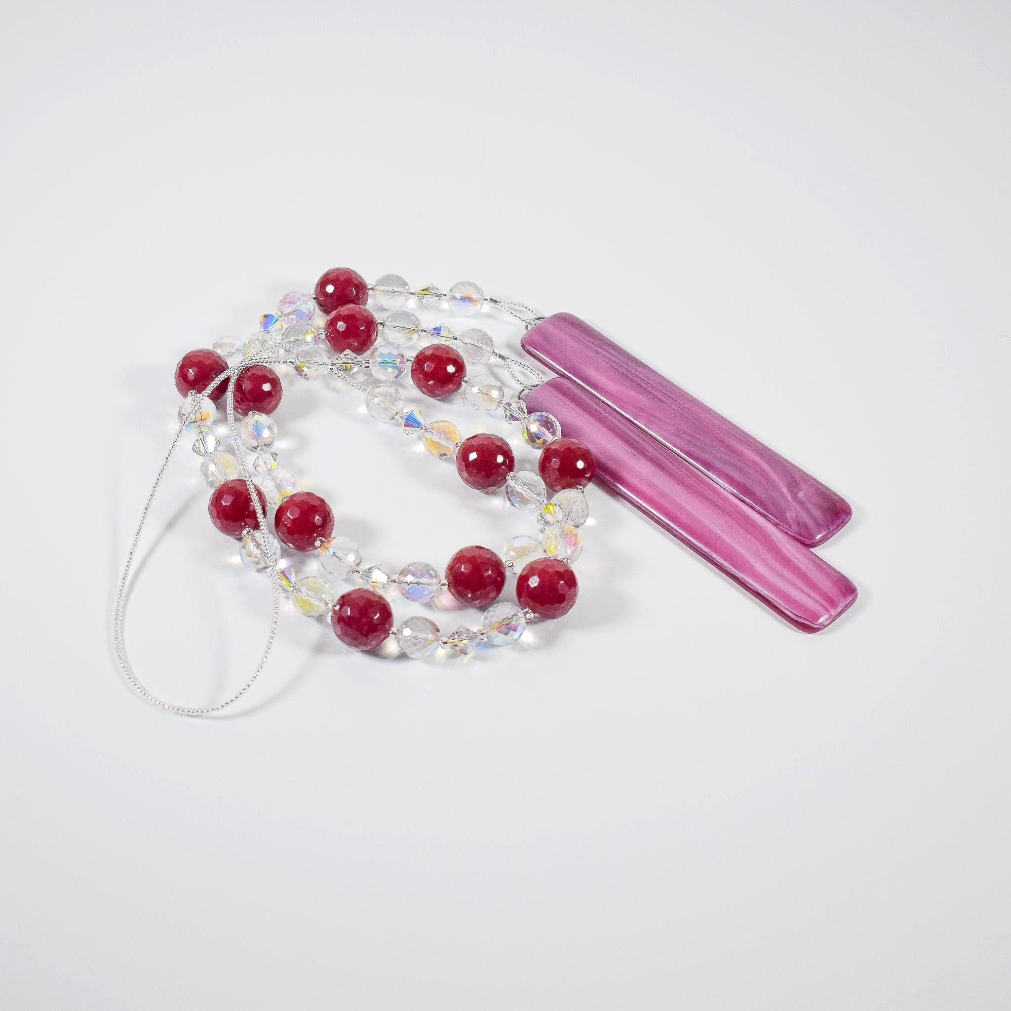 Large cranberry red jade stones beaded with glass and crystals, anchored by two pieces of dark cranberry pink fused glass, coiled on flat white surface.