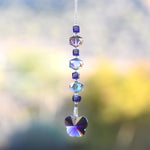 A handmade sun catcher featuring purple and iridescent crystal beads, finished with a faceted crystal butterfly pendant that reflects light in purple, blue, and gold tones. Suspended from a wire covered in silver-lined seed beads against a blurred outdoor background.