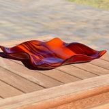 Large square glass bowl with wavy edges and a square center foot, viewed from the side sitting on a slatted wood table outside in the sun.