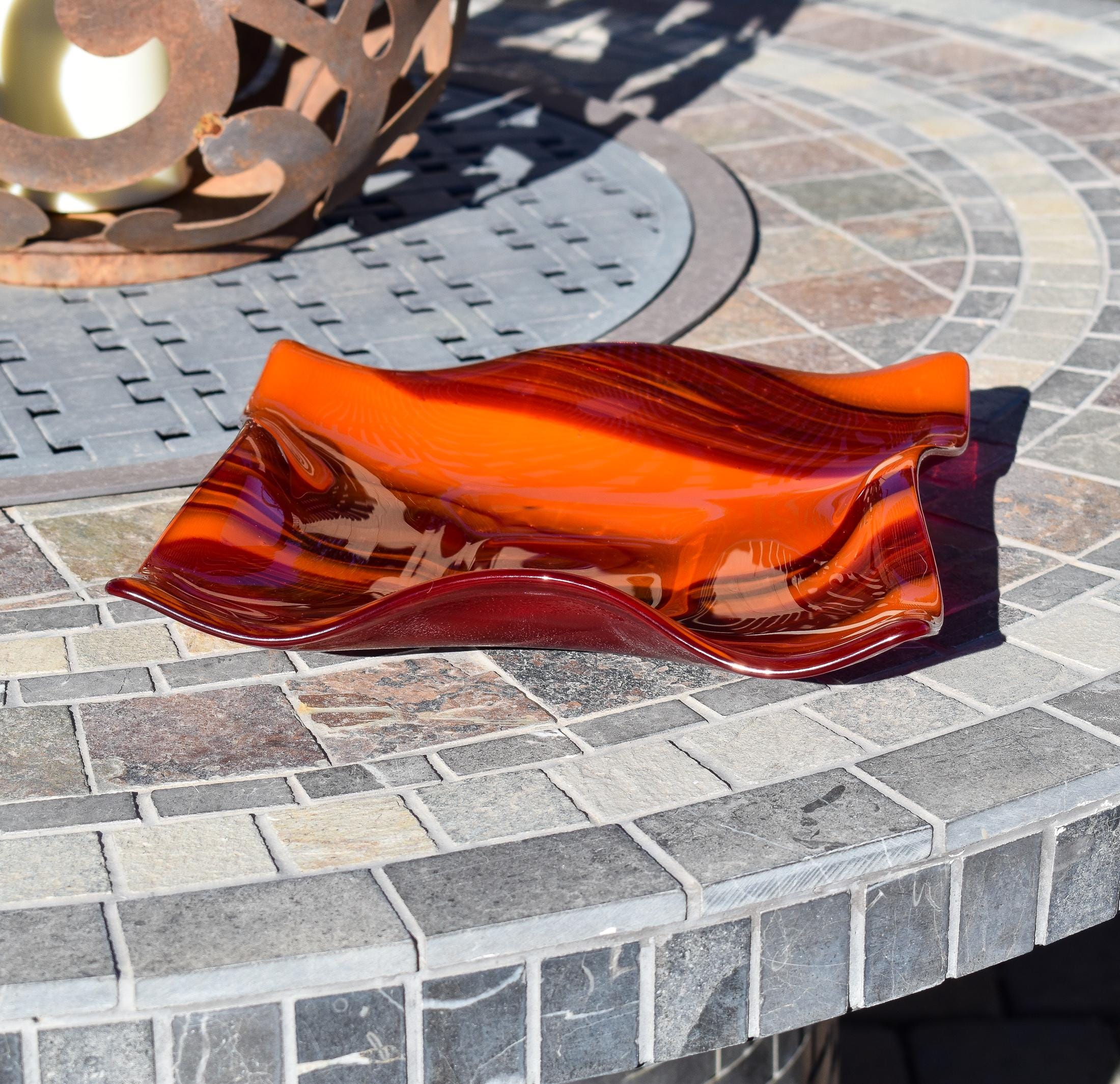 Large square glass bowl with wavy edges and a square center foot, viewed at an angle sitting on a stone table.