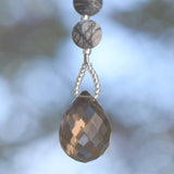 A faceted, teardrop-shaped smoky quartz gemstone strung on wire and hanging against blurred background. Above the quartz are two round, patterned beads in shades of gray and cream.