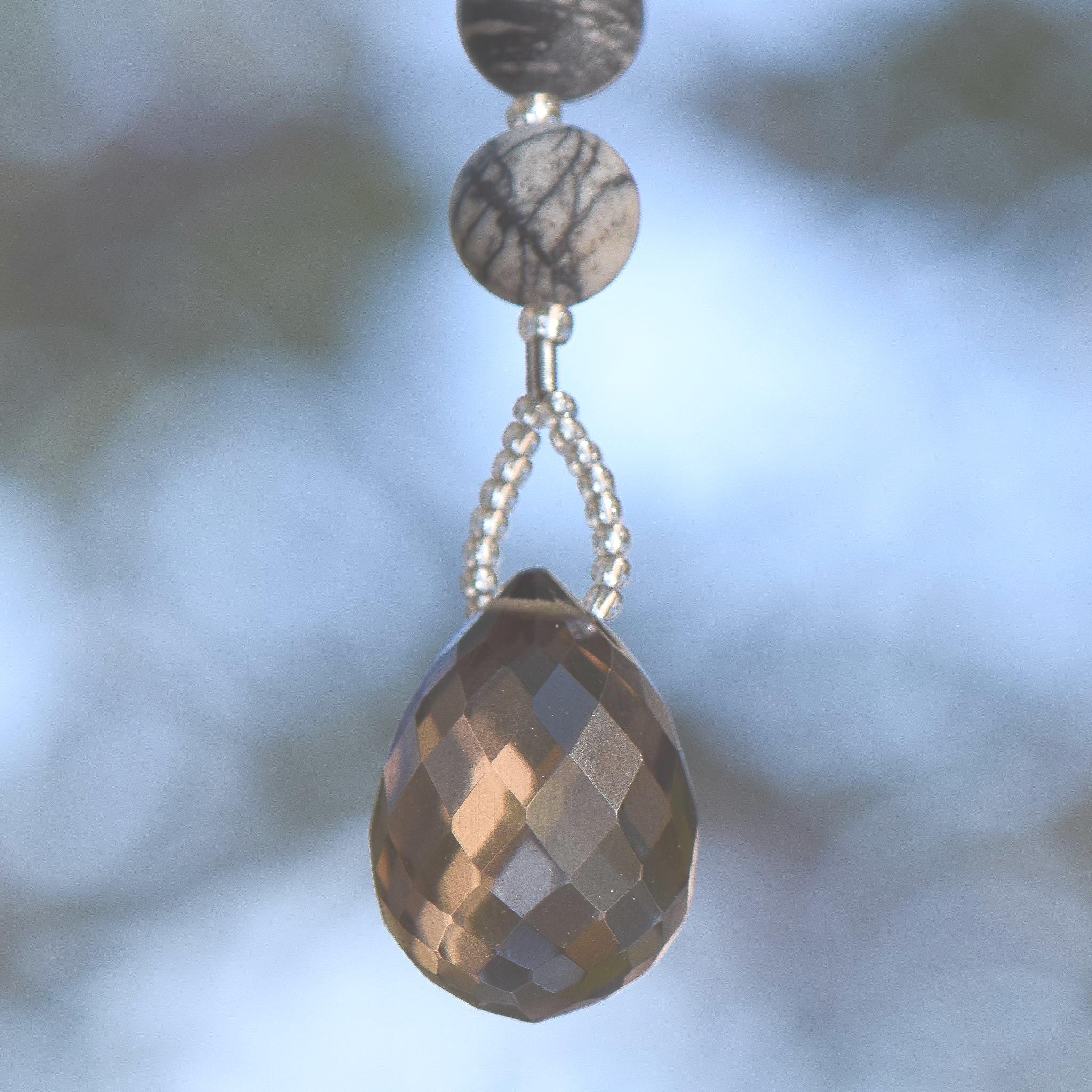 A faceted, teardrop-shaped smoky quartz gemstone strung on wire and hanging against blurred background. Above the quartz are two round, patterned beads in shades of gray and cream.
