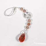 A crystal beaded car charm with a teardrop-shaped pink crystal at the bottom. The charm is made of clear and light pink crystals and is attached to a wire covered in seed beads. The sun catcher is coiled on a white surface.