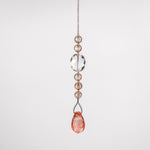 A crystal beaded car charm with a teardrop-shaped pink crystal at the bottom. The charm is made of clear and light pink crystals and is attached to a wire covered in seed beads. The sun catcher is hanging in front of a white surface.