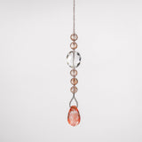 A crystal beaded car charm with a teardrop-shaped pink crystal at the bottom. The charm is made of clear and light pink crystals and is attached to a wire covered in seed beads. The sun catcher is hanging in front of a white surface.
