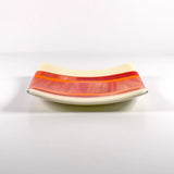 Square fused glass tray with vanilla cream colored base and a striped and streaky design of red, orange and white glass. Tray is 6" square with upturned corners. Sitting on a white surface.