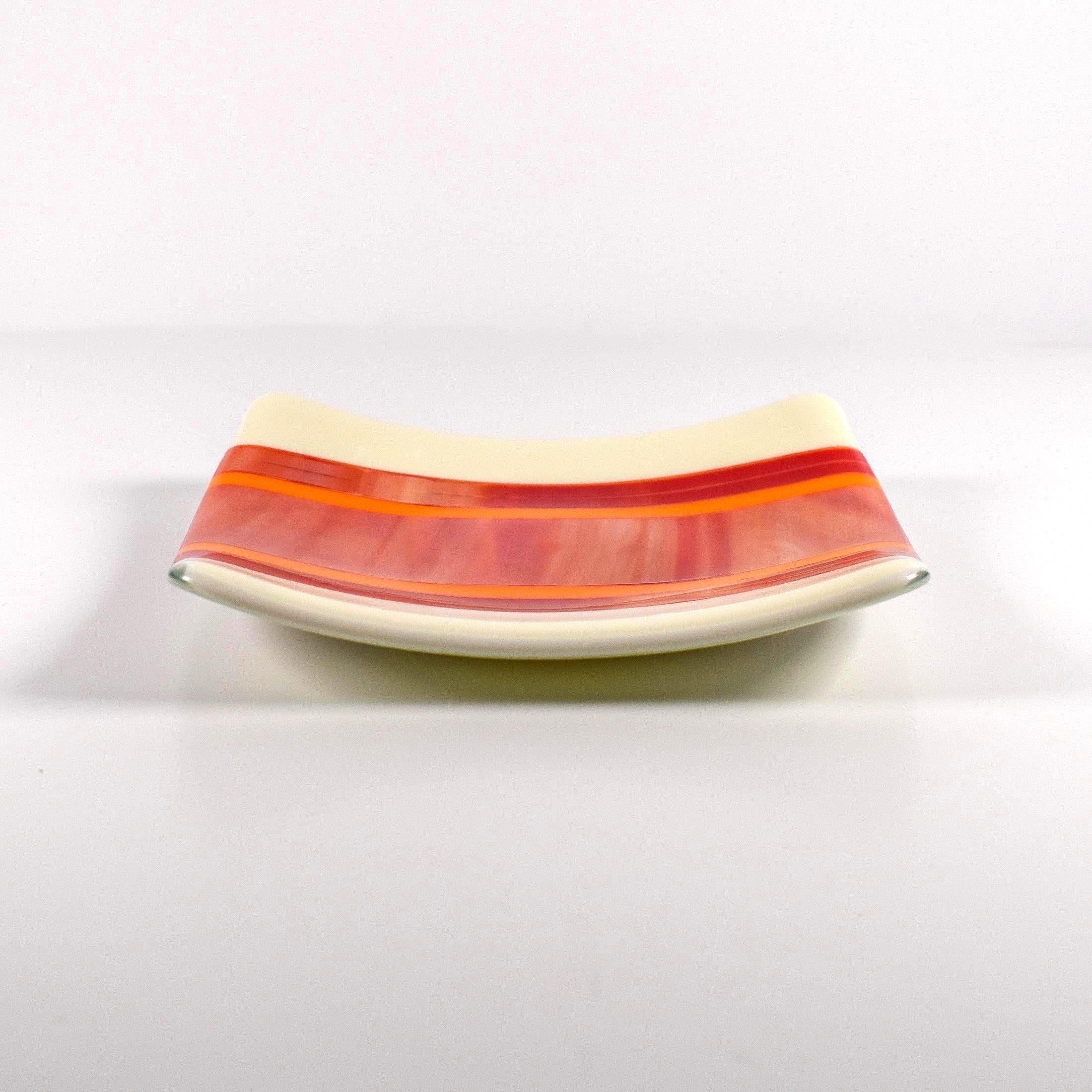 Square fused glass tray with vanilla cream colored base and a striped and streaky design of red, orange and white glass. Tray is 6" square with upturned corners. Sitting on a white surface.