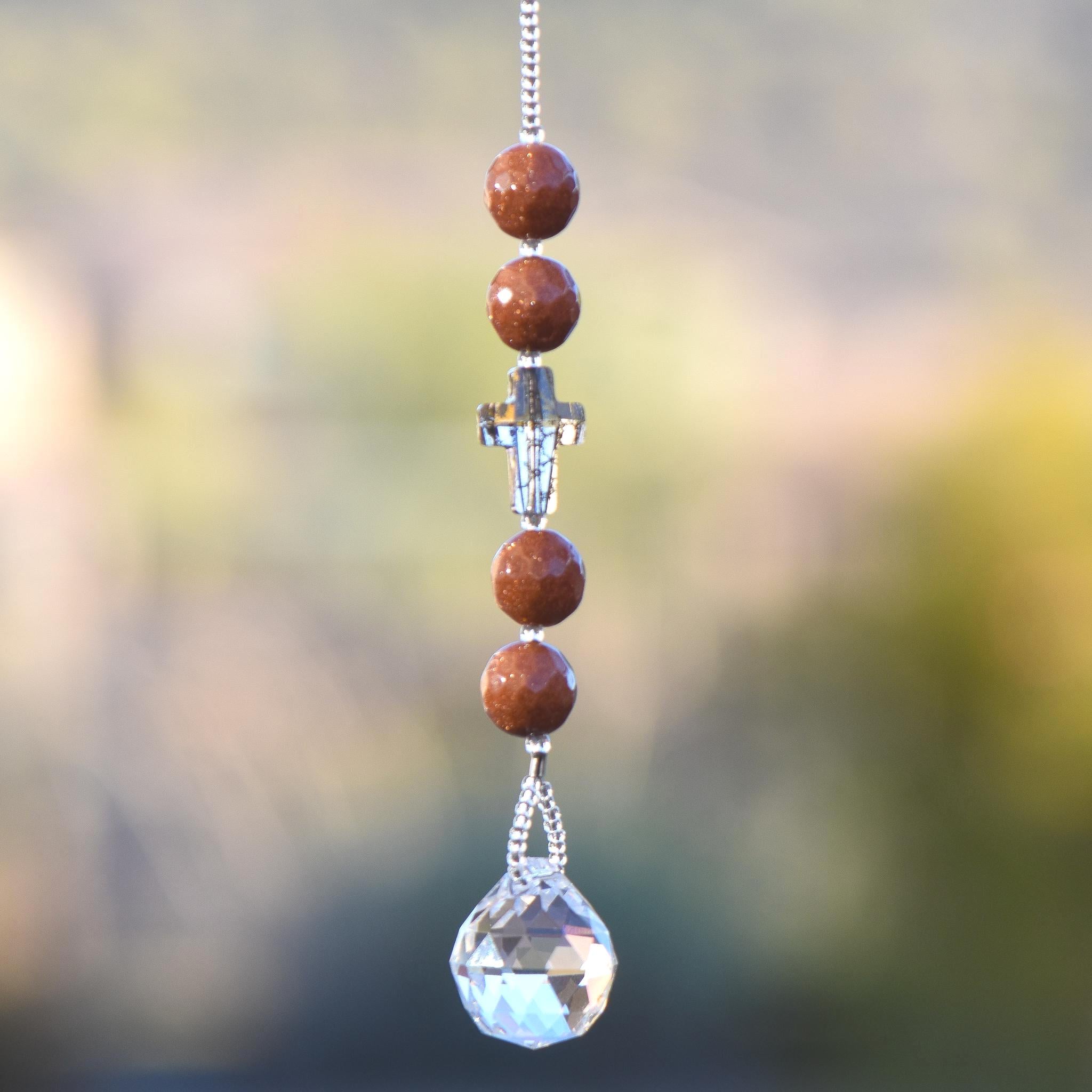 Four round goldstone beads flanking crystal cross bead in gold patina, anchored by small crystal prism, hanging vertically against blurred background.