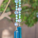 Large faceted glass crystal beads strung on wire with smaller howlite stone beads, hanging vertically from tree, anchored by two pieces of kiln formed glass in transparent sky blue. Against blurred tree background.