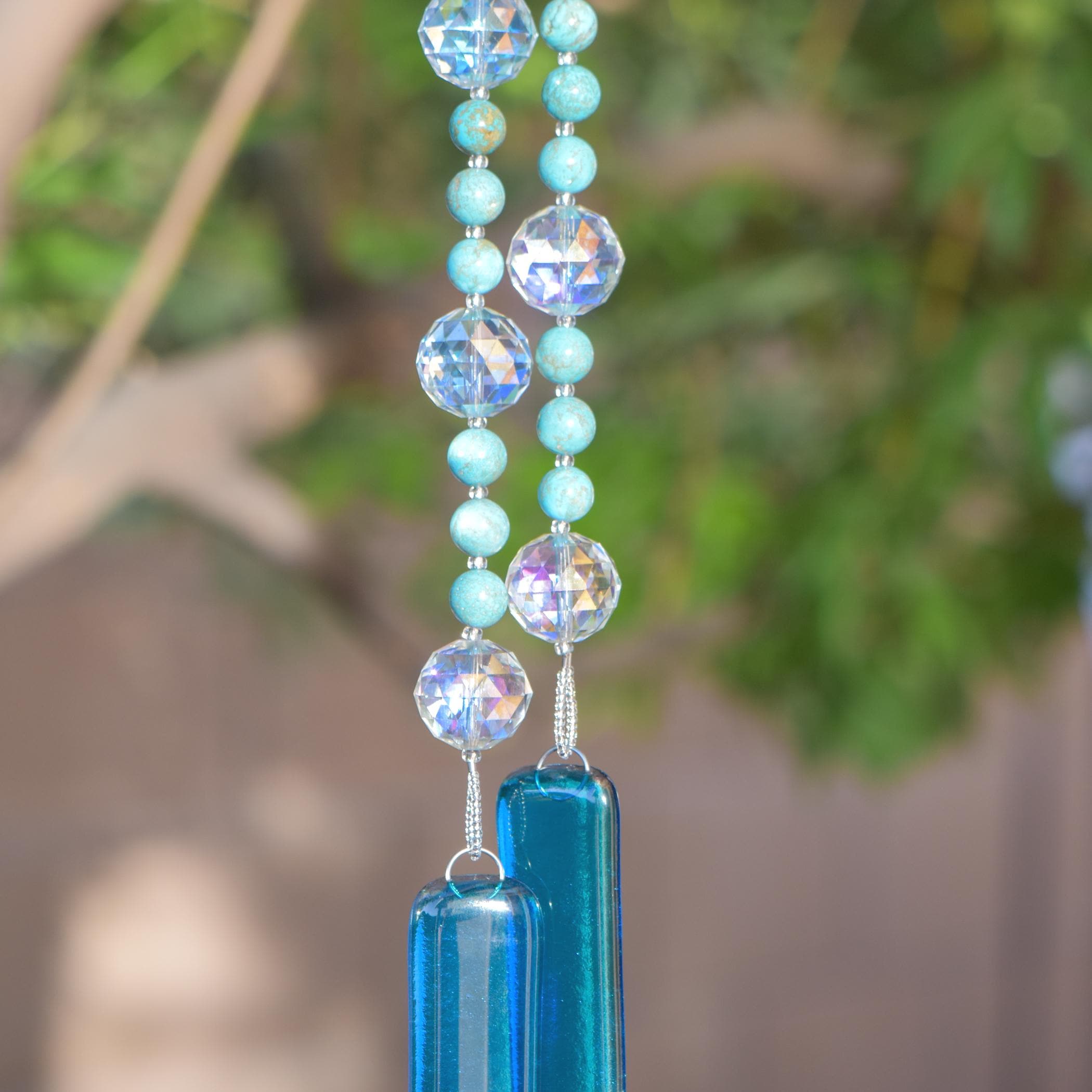 Large faceted glass crystal beads strung on wire with smaller howlite stone beads, hanging vertically from tree, anchored by two pieces of kiln formed glass in transparent sky blue. Against blurred tree background.